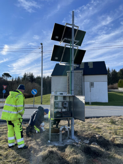 Två arbetare i synliga jackor som inspekterar och underhåller en elektronisk LEAKCELL LC221 vid vägkanten nära en solcellspanel.