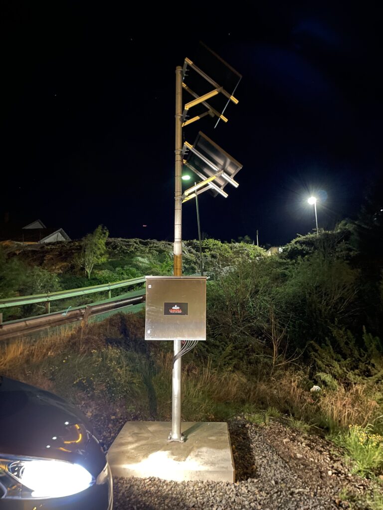En metallstolpe med flera riktade antenner och en kontrollbox, upplyst av gatlyktor på natten, med vegetation och en bil synlig i bakgrunden.